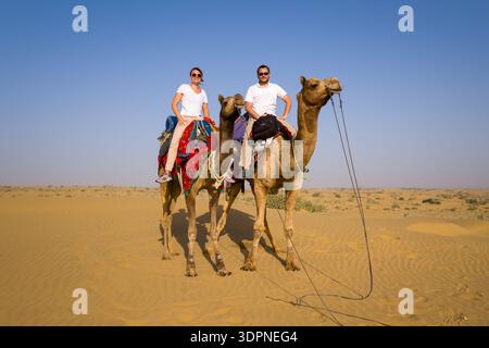 Jaisalmer, Inde - 29 mars 2022 : deux personnes montent à dos de chameaux avec des selles colorées dans le paysage sablonneux du désert du Thar sous un ciel dégagé. Banque D'Images