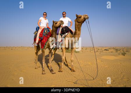 Jaisalmer, Inde - 29 mars 2022 : deux personnes montent à dos de chameaux avec des selles colorées dans le paysage sablonneux du désert du Thar sous un ciel dégagé. Banque D'Images