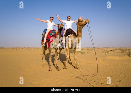 Jaisalmer, Inde - 29 mars 2022 : deux personnes montent à dos de chameaux avec des selles colorées, levant les bras, dans le paysage sablonneux du désert du Thar sous un ciel dégagé. Banque D'Images