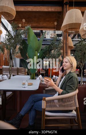 Jeune femme ayant un appel téléphonique et buvant du café à la terrasse du café Banque D'Images