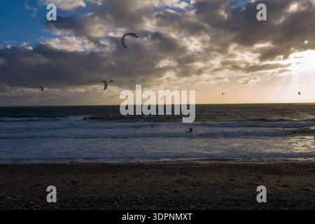 Le Havre, France - 25 août 2020 : plusieurs kitesurfeurs chevauchent les vagues sur la mer avec des cerfs-volants colorés dans les airs, sous un ciel nuageux dramatique au coucher du soleil, avec une plage de galets au premier plan. Banque D'Images