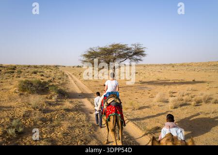 Jaisalmer, Inde - 29 mars 2022 : L'image montre des gens chevauchant des chameaux le long d'une piste de terre à travers un terrain aride avec une végétation clairsemée et un seul arbre dans le désert du Thar près de Jaisalmer. Banque D'Images