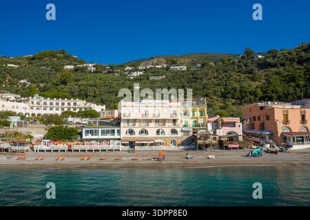 Marina del Cantone, Italie - 6 octobre 2020 : L'image montre une rangée de bâtiments en bord de mer et d'hôtels le long de la plage de galets de Marina del Cantone, avec des collines couvertes de verdure s'élevant en arrière-plan sous un ciel bleu clair. Banque D'Images