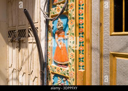 Udaipur, Inde - 23 mars 2022 : une vue rapprochée d'un cadre de porte en bois sculpté et peint avec une figure traditionnelle Rajasthani en tenue orange, des motifs floraux et des portes métalliques adjacentes, avec des câbles électriques visibles à la lumière du soleil. Banque D'Images