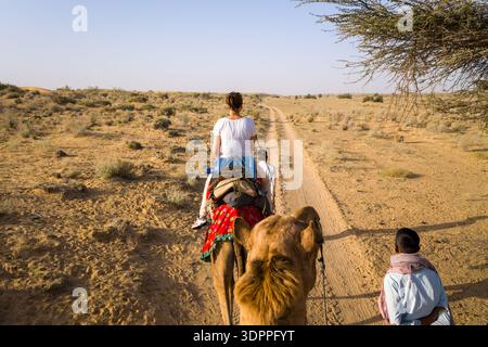 Jaisalmer, Inde - 29 mars 2022 : L'image montre des gens chevauchant des chameaux le long d'une piste de terre sablonneuse à travers un terrain aride avec une végétation clairsemée et une branche d'arbre au-dessus du ciel dans le désert de Thar près de Jaisalmer. Banque D'Images