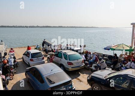 Phnom Penh, Cambodge - 7 février 2020 : voitures, motos et passagers sont bondés sur le pont d'un ferry traversant le Mékong, avec des gens assis et debout près de leurs véhicules sous un ciel dégagé et la rive opposée visible au loin. Banque D'Images