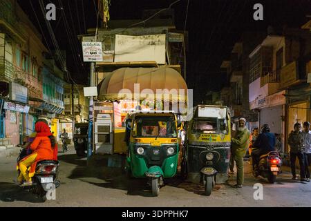 Jodhpur, Inde - 25 mars 2022 : L'image montre une scène de rue nocturne avec deux pousse-pousse automobiles garés côte à côte devant un magasin avec un auvent jaune, une femme en rouge à cheval sur un scooter, plusieurs personnes debout et assises sur des scooters, et des fils électriques aériens visibles parmi les bâtiments de la vieille ville. Banque D'Images