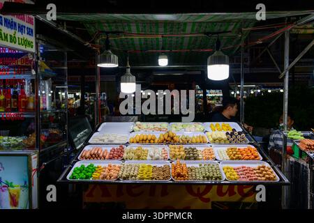 CAN Tho, Vietnam - 19 janvier 2020 : des plateaux de brochettes assorties de nourriture de rue, y compris de la viande, des fruits de mer et des légumes, sont exposés sous des lumières vives sur un stand de marché nocturne, avec deux vendeurs visibles derrière le comptoir et une signalisation lumineuse en arrière-plan. Banque D'Images