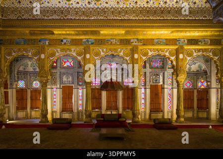 Jodhpur, Inde - 25 mars 2022 : L'image montre une salle de réception richement décorée au fort de Mehrangarh avec des arches dorées, des vitraux, des panneaux muraux peints et des sièges traditionnels avec des coussins et une verrière cérémonielle. Banque D'Images