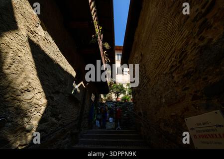 Isola Bella, Italie - 30 septembre 2020 : une étroite ruelle en pierre avec un escalier est visible entre de vieux bâtiments avec des balcons en bois, des plantes en pot, et un panneau pour il Fornello Bottega & Cucina sous un ciel bleu clair. Banque D'Images