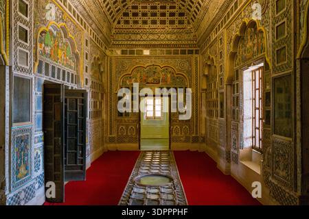 Jodhpur, Inde - 25 mars 2022 : L'image montre une pièce richement décorée au fort de Mehrangarh avec des miroirs complexes, des peintures murales colorées, des portes voûtées et des fenêtres avec des volets en bois, avec un tapis rouge couvrant le sol. Banque D'Images