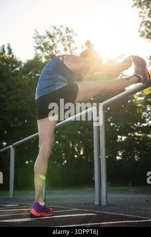 Femme étire la jambe sur les balustrades entourées d'arbres et de lumière du soleil, coureuse de fond moyenne Katharina Jaiser, Gechingen, Allemagne Banque D'Images