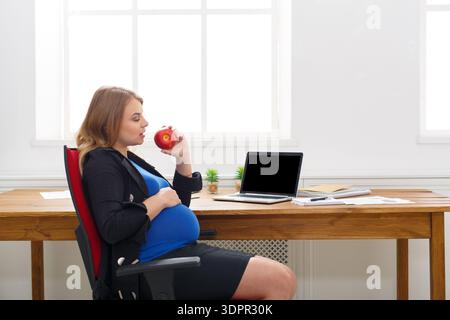 Femme enceinte est assis au bureau dans le bureau manger Apple tout en travaillant sur ordinateur portable Banque D'Images