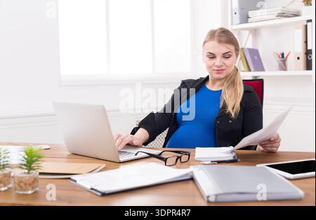 Femme enceinte travaillant sur un ordinateur portable tout en examinant des documents dans un bureau Banque D'Images