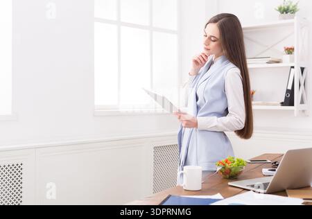 Femme d'affaires examine la paperasse tout en restant debout dans le bureau moderne pendant les heures de jour Banque D'Images