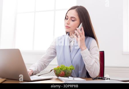 Femme travaillant au bureau tout en parlant au téléphone et en mangeant de la salade pendant les heures de milieu de matinée Banque D'Images