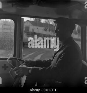 Région de Donetsk, Ukraine, URSS. 1960s-1970s. Portrait d'un conducteur au volant d'un véhicule soviétique de premiers secours ou d'ambulance au Donrybkombinat dans le nord du Donbass. L'homme, portant une casquette et une veste, semble concentré lorsqu'il conduit le véhicule. Une croix rouge est visible sur le panneau intérieur de porte. En arrière-plan, à travers la fenêtre, d'autres camions soviétiques et un groupe de travailleurs sont visibles dans un cadre rural. Cette scène historique documente la sécurité au travail et le soutien médical dans une grande entreprise soviétique dans le Donbass pacifique. Banque D'Images