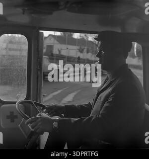 Région de Donetsk, Ukraine, URSS. 1960s-1970s. Portrait d'un conducteur au volant d'un véhicule soviétique de premiers secours ou d'ambulance au Donrybkombinat dans le nord du Donbass. L'homme, portant une casquette et une veste, semble concentré lorsqu'il conduit le véhicule. Une croix rouge est visible sur le panneau intérieur de porte. En arrière-plan, à travers la fenêtre, d'autres camions soviétiques et un groupe de travailleurs sont visibles dans un cadre rural. Cette scène historique documente la sécurité au travail et le soutien médical dans une grande entreprise soviétique dans le Donbass pacifique. Banque D'Images