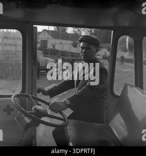 Région de Donetsk, Ukraine, URSS. 1960s-1970s. Portrait précis d'un conducteur derrière le volant d'un véhicule de premiers soins au Donrybkombinat dans le nord du Donbass. L'homme porte une casquette plate traditionnelle et une veste. À travers les fenêtres, un camion soviétique et un groupe de personnes sont visibles sur le territoire de l'entreprise. Le symbole de croix rouge sur la porte indique la destination médicale du transport. Cette scène historique documente la sécurité au travail et l'infrastructure sociale de l'ère soviétique dans la région pacifique du Donbass avant la guerre. Banque D'Images