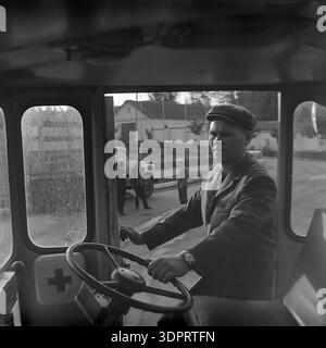 Région de Donetsk, Ukraine, URSS. 1960s-1970s. Portrait précis d'un conducteur derrière le volant d'un véhicule de premiers soins au Donrybkombinat dans le nord du Donbass. L'homme porte une casquette plate traditionnelle et une veste. À travers les fenêtres, un camion soviétique et un groupe de personnes sont visibles sur le territoire de l'entreprise. Le symbole de croix rouge sur la porte indique la destination médicale du transport. Cette scène historique documente la sécurité au travail et l'infrastructure sociale de l'ère soviétique dans la région pacifique du Donbass avant la guerre. Banque D'Images