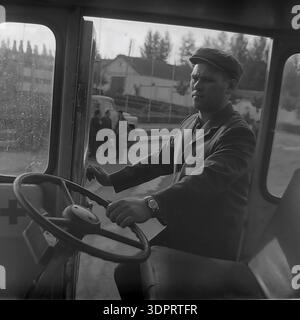 Région de Donetsk, Ukraine, URSS. 1960s-1970s. Portrait précis d'un conducteur derrière le volant d'un véhicule de premiers soins au Donrybkombinat dans le nord du Donbass. L'homme porte une casquette plate traditionnelle et une veste. À travers les fenêtres, un camion soviétique et un groupe de personnes sont visibles sur le territoire de l'entreprise. Le symbole de croix rouge sur la porte indique la destination médicale du transport. Cette scène historique documente la sécurité au travail et l'infrastructure sociale de l'ère soviétique dans la région pacifique du Donbass avant la guerre. Banque D'Images