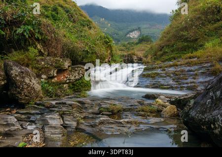 Une petite cascade coule sur la roche en couches dans un ruisseau peu profond entouré d'une végétation dense dans une vallée montagneuse du nord du Vietnam. Banque D'Images