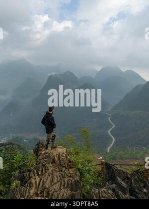 Une vue aérienne oblique montre une personne debout sur un point de vue rocheux surplombant une route de montagne sinueuse et des collines karstiques boisées dans le nord du Vietnam. Banque D'Images