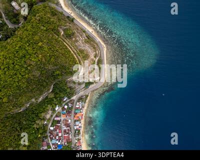 Une petite ville côtière longe le rivage à côté d'une route côtière sinueuse, vue directement d'en haut avec une eau bleue claire et des collines boisées à l'intérieur des terres. Banque D'Images