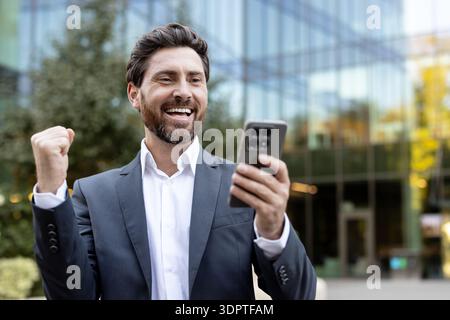 Heureux jeune homme d'affaires barbu habillé en vêtements formels debout à l'extérieur. Célébrant un geste de triomphe en regardant son smartphone avec un grand sourire. Représentant le succès en ligne et les nouvelles positives Banque D'Images