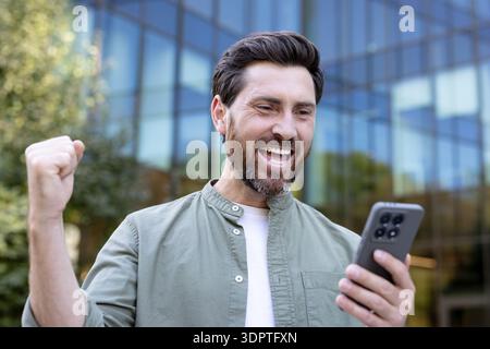 Homme souriant se sentant excité et triomphant, recevant d'excellentes nouvelles sur son téléphone portable avec un poing serré, célébrant la victoire et la réussite en plein air Banque D'Images