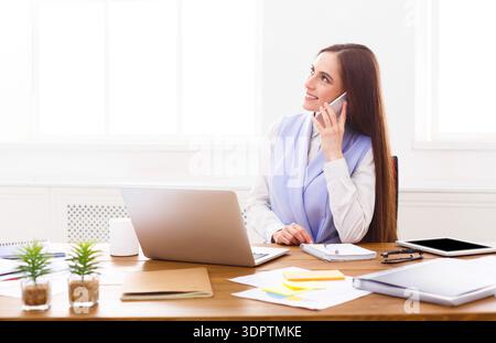 Femme d'affaires parle sur smartphone tout en étant assis au bureau dans le bureau pendant la journée Banque D'Images