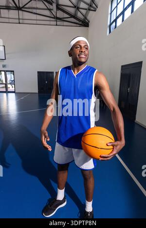 Homme afro-américain portant un maillot bleu tenant le basket-ball sur le terrain par le backboard à l'intérieur de la salle de gym Banque D'Images