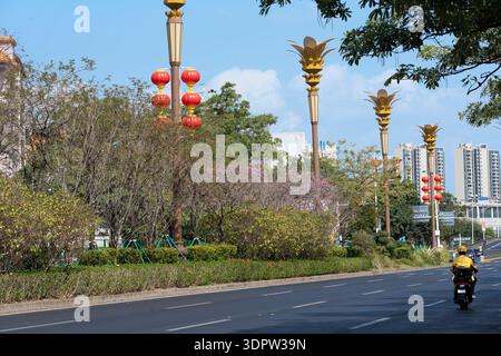 Scène de rue à Guangzhou avec lampadaires dorés, lanternes rouges, motocycliste, bâtiments modernes et arbres fleuris sous ciel clair Banque D'Images