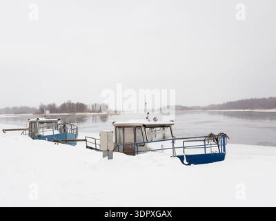 Les bateaux de pêche enneigés reposent tranquillement le long d'une rivière gelée sous un ciel gris hivernal. Une scène hivernale tranquille dans un petit port de pêche. Banque D'Images