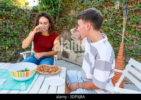 Jeune femme et homme transgenre buvant de l'eau ensemble sur une terrasse extérieure ensoleillée lors d'un rassemblement décontracté. Mise en valeur des moments de vie authentiques Banque D'Images