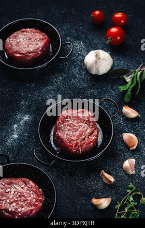Un gros plan de steaks de filet mignon crus assaisonnés de sel et associés à des gousses d'ail fraîches, des tomates cerises et des herbes sur un dos à texture foncée Banque D'Images