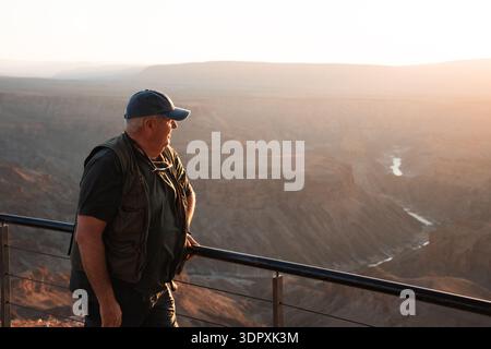 Une personne se tient debout sur une balustrade, profitant de la vue imprenable sur Fish River Canyon en Namibie au coucher du soleil. Le soleil doré jette des teintes chaudes sur th Banque D'Images