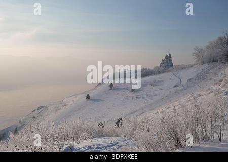 Paysage hivernal captivant avec des arbres givrés et une colline enneigée sereine, avec un bâtiment religieux lointain émergeant à travers la brume sous-jacente Banque D'Images