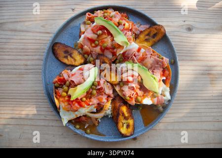 Un gros plan des Huevos Motuleños traditionnels du Yucatan servis avec du riz et des bananes frites, des œufs et de l'avocat sur une assiette texturée à Sisal, Yucatan, Mexique. Banque D'Images