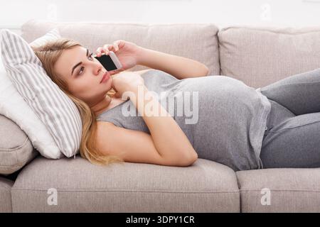 Femme enceinte allongée sur canapé et parlant au téléphone dans le salon pendant la journée Banque D'Images