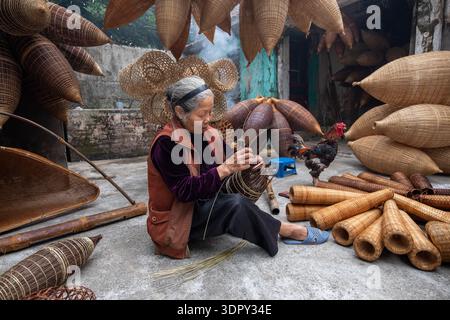 Hưng Yên, Vietnam - 15 décembre 2019 : vue d'un artisan tissant des pièges à poissons en bambou complexes, entouré par son artisanat, alors qu'un coq se dresse à proximité, le Banque D'Images