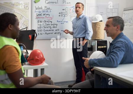 Homme d'âge moyen caucasien debout près du tableau blanc expliquant le calendrier du projet au jeune homme adulte noir et homme d'âge moyen caucasien dans un cadre de bureau, casques de construction sur la table Banque D'Images
