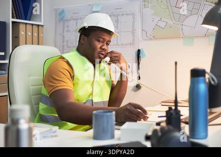 Homme noir d'âge moyen portant un gilet de sécurité et un casque de sécurité assis au bureau parlant sur un téléphone fixe, tenant un stylo examinant des documents avec des plans architecturaux sur le mur Banque D'Images