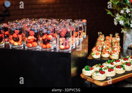 Assortiment vibrant de coupes de fruits à baies en couches dans des verres transparents accompagnés de délicieux canapés au fromage à la crème avec du caviar rouge et des herbes sur un plateau en ardoise noire Banque D'Images