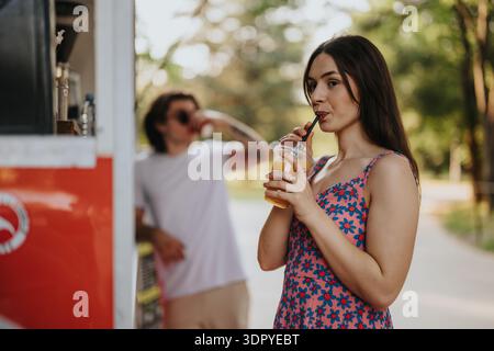 Femme profitant d'une boisson rafraîchissante de jus dans un parc ensoleillé à côté d'un camion de nourriture rouge Banque D'Images