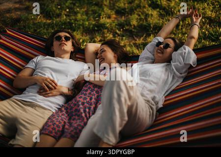 Trois amis se relaxant sur un hamac rayé à l'extérieur, souriant et profitant d'un moment ensoleillé et insouciant Banque D'Images