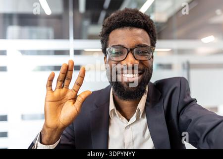 Jeune homme d'affaires africain en lunettes et veste de costume souriant largement et agitant à la caméra lors d'une réunion virtuelle ou d'un appel vidéo, prise de vue professionnelle conviviale et accessible Banque D'Images