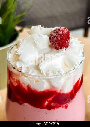 Milk-Shake aux fraises garni de crème fouettée et de framboise servi dans un verre à la table du café en gros plan. Boisson sucrée, dessert, rafraîchissement, style de vie Banque D'Images