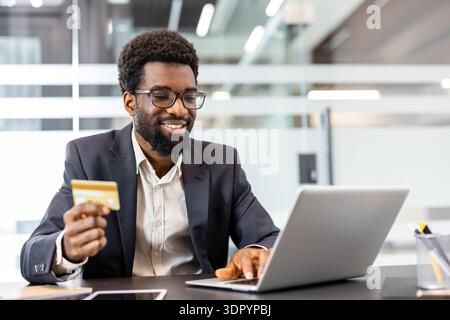 Homme souriant portant un costume et des lunettes, assis à un bureau, tenant une carte de crédit dorée et tapant sur un ordinateur portable, profitant du shopping numérique sécurisé Banque D'Images