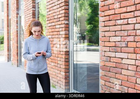 Jeune femme marche sur le trottoir tout en utilisant smartphone près de Brick Wall Banque D'Images